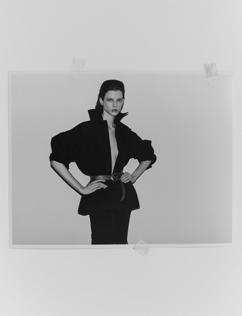 Monochrome fashion portrait of model Sandra Murray wearing a belted black jacket with hands on hips against a pale studio backdrop.