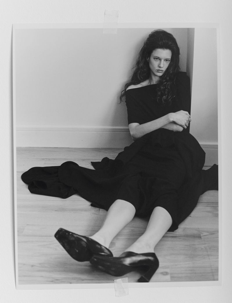 Black and white photograph of Sandra Murray seated on a wooden floor in a black dress and heeled mules, leaning against a wall.