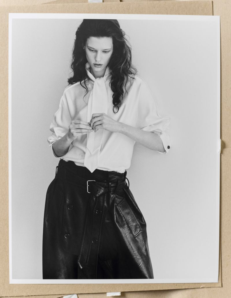 Sandra Murray wearing a white tie-neck blouse and belted black leather skirt in a monochrome studio portrait.