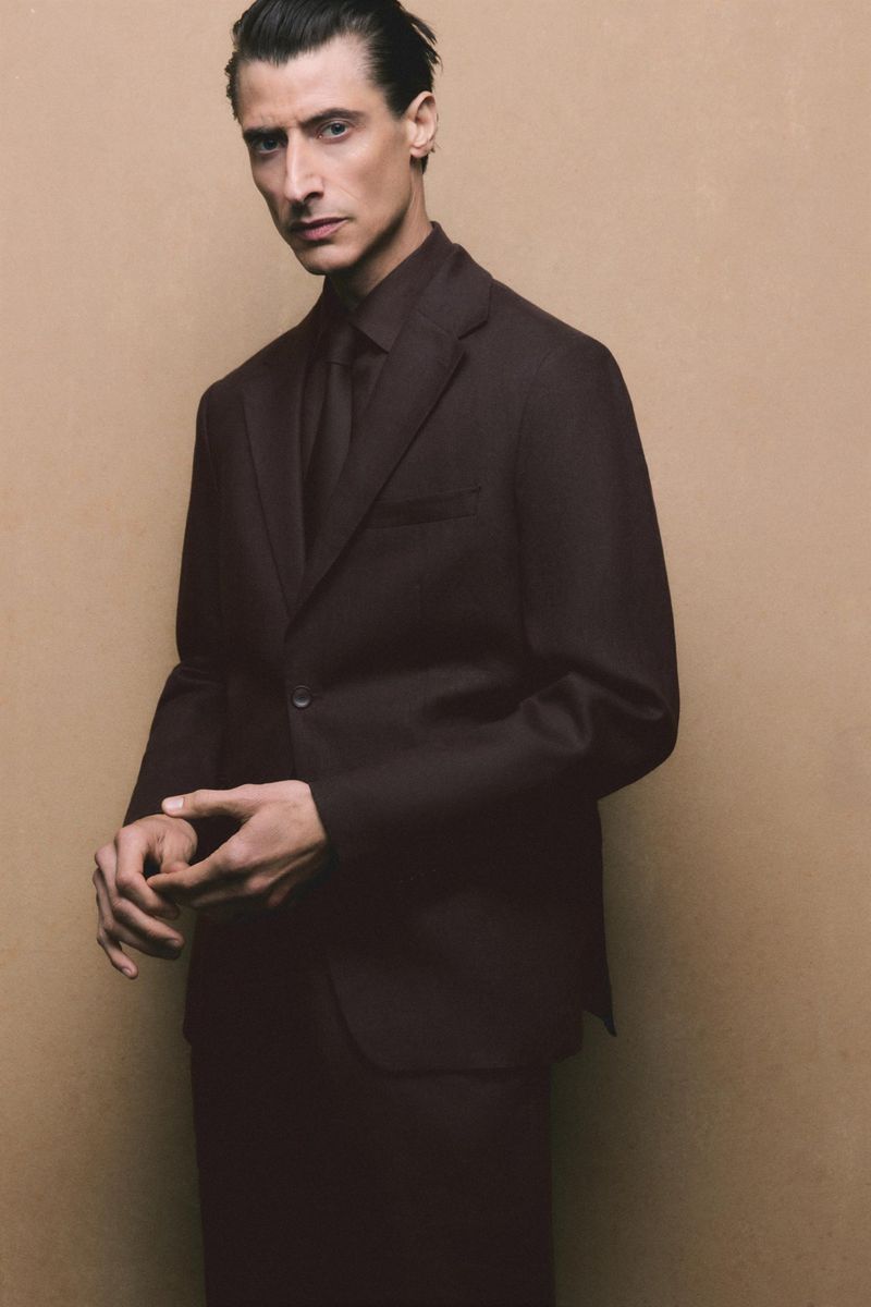 Man in a dark suit and tie standing in front of a tan background, fashion portrait, male formalwear, dark color palette, menswear.