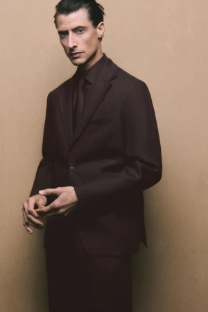 Man in a dark suit and tie standing in front of a tan background, fashion portrait, male formalwear, dark color palette, menswear.