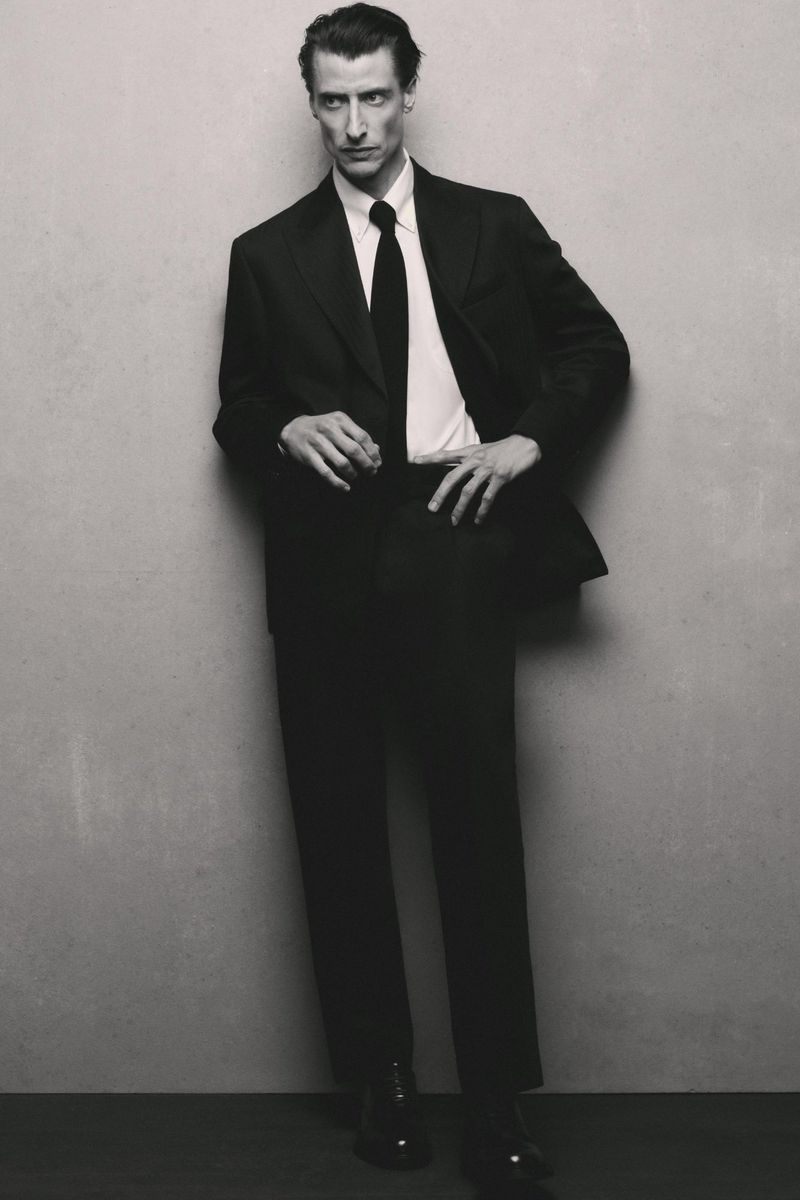 Model Jonas Mason posing in a tailored black suit with a white shirt and black tie against a grey textured studio backdrop.