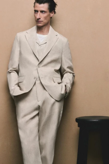A man in a tan linen suit and white undershirt leans against a tan wall next to a black stool in a studio portrait.