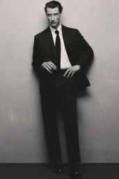 Model Jonas Mason posing in a tailored black suit with a white shirt and black tie against a grey textured studio backdrop.