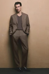 Model Jonas Mason standing in a brown tailored suit with hands in pockets against a warm taupe studio backdrop.