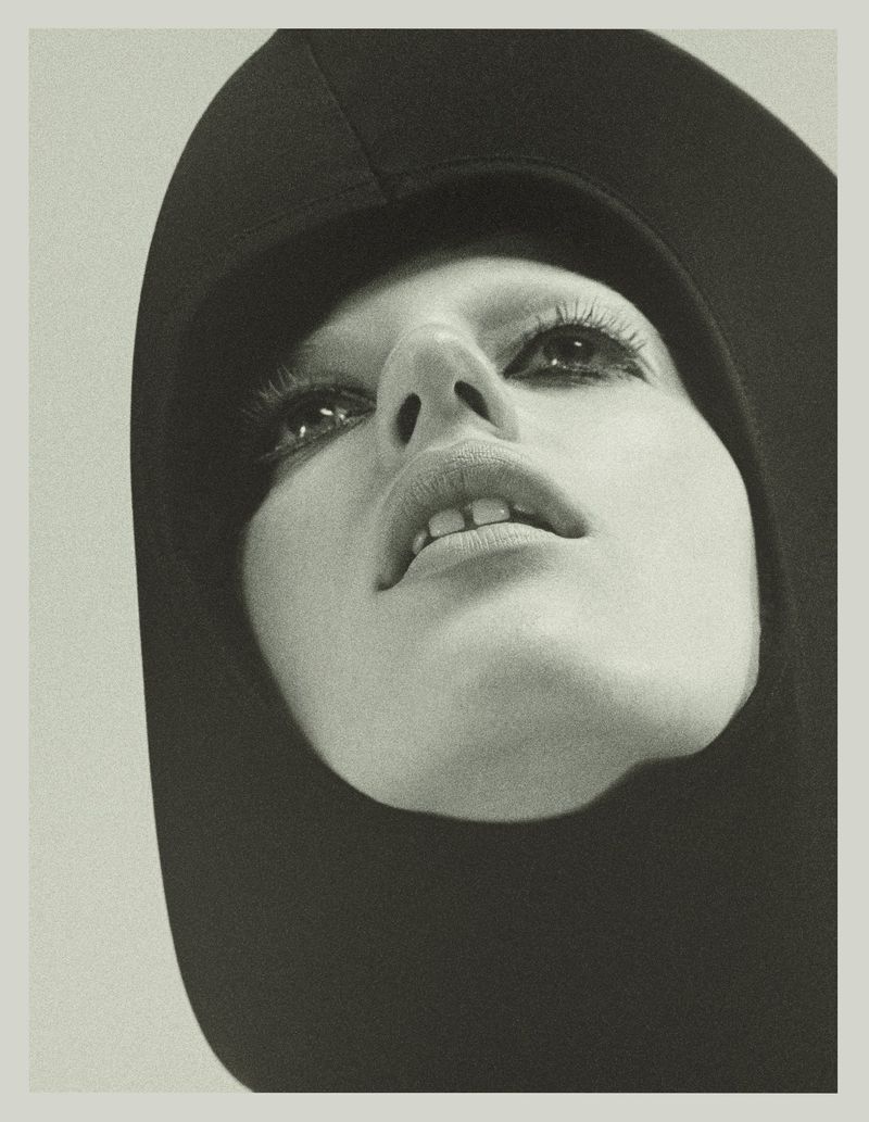 Close-up portrait of model Julia Banas wearing a black hooded garment with dark eye makeup against a light background.
