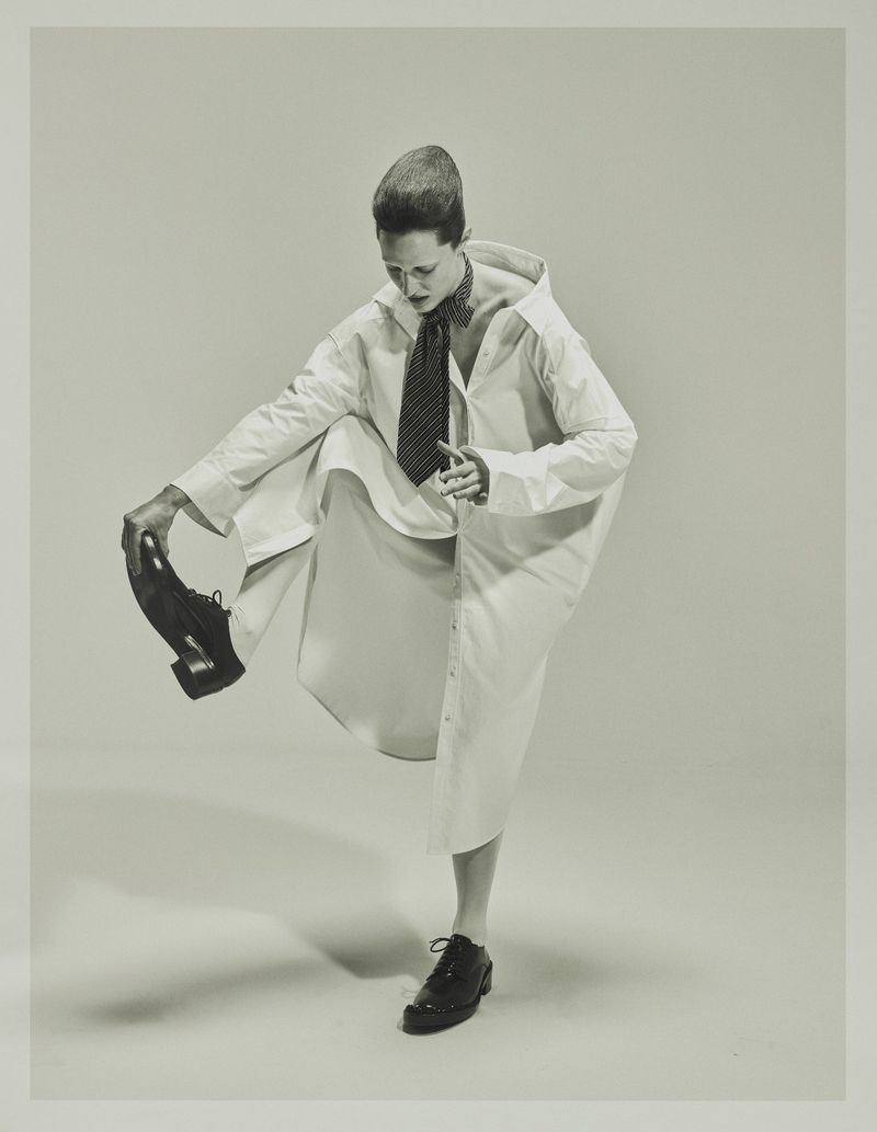 Model Julia Banas in an oversized white shirt dress and necktie balancing on one leg against a pale studio backdrop.