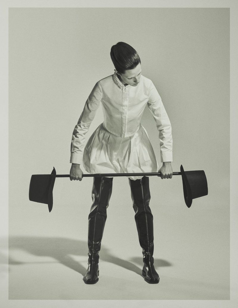 Julia Banas posing with a top hat barbell wearing a white balloon skirt and black thigh-high boots against a grey studio backdrop.