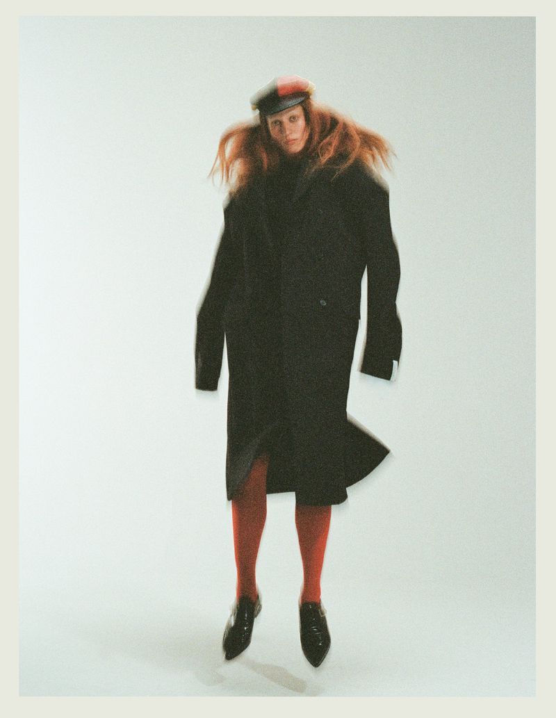 Model in long black coat and red tights jumping against a plain white background for Tidal Magazine