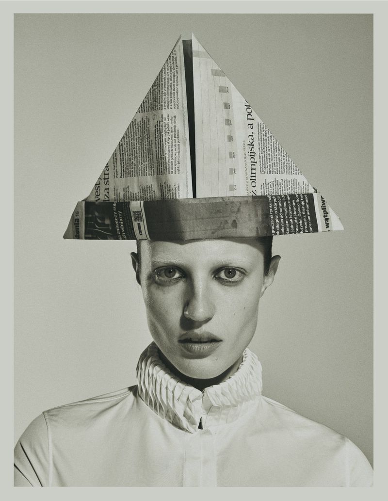 Model Julia Banas wears an origami newspaper hat and a ruffled high-collar shirt against a pale studio background.