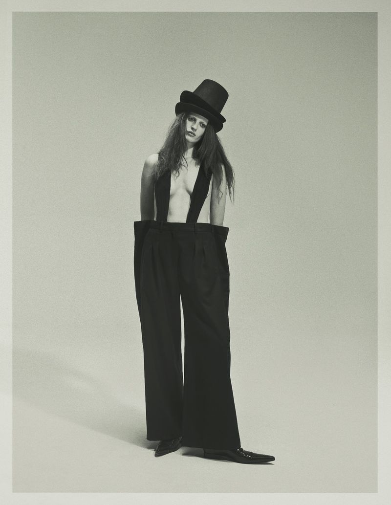 Model Julia Banas posing in oversized black trousers with suspenders and a black top hat against a neutral studio backdrop.