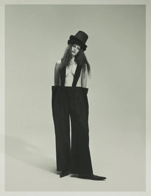 Model Julia Banas posing in oversized black trousers with suspenders and a black top hat against a neutral studio backdrop.