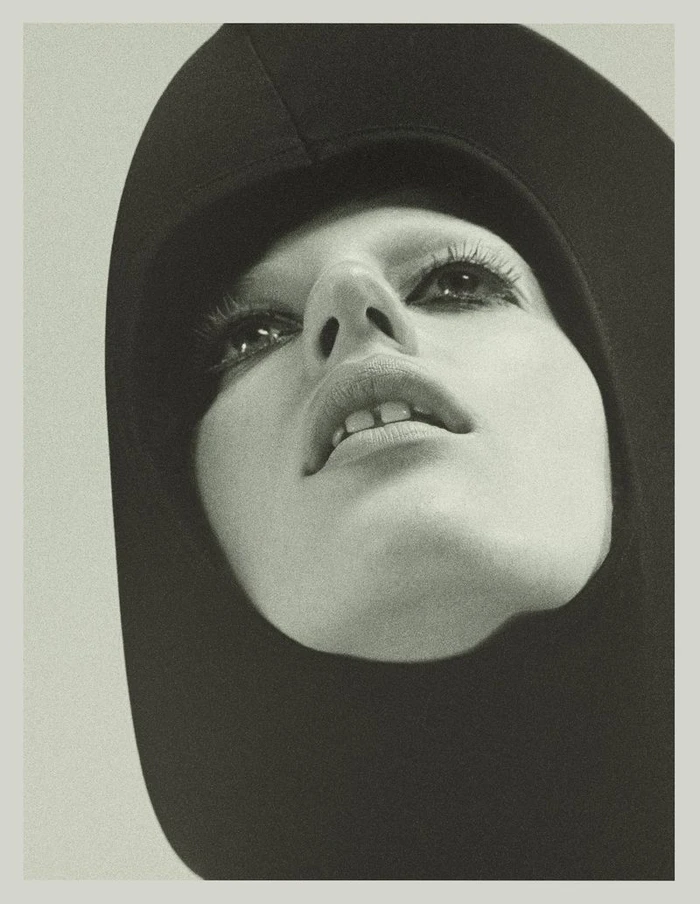 Close-up portrait of model Julia Banas wearing a black hooded garment with dark eye makeup against a light background.