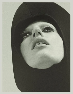 Close-up portrait of model Julia Banas wearing a black hooded garment with dark eye makeup against a light background.