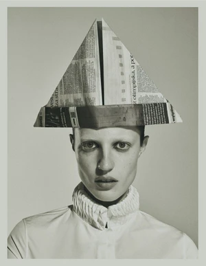 Model Julia Banas wears an origami newspaper hat and a ruffled high-collar shirt against a pale studio background.