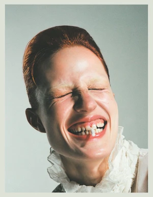 Julia Banas laughing with eyes closed wearing a white ruffled collar against a pale grey studio backdrop.