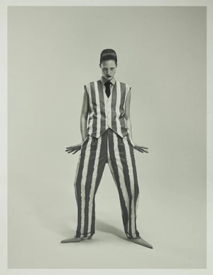 Model Julia Banas posing in a vertical striped vest and matching wide-leg trousers with a black tie against a seamless studio background.