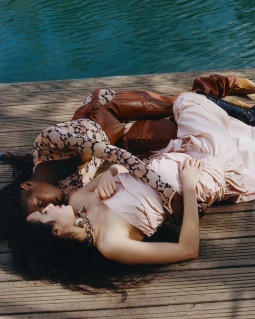 Two women lie together on a wooden dock alongside blue water. One woman wears a pink dress, while the other wears a patterned top.
