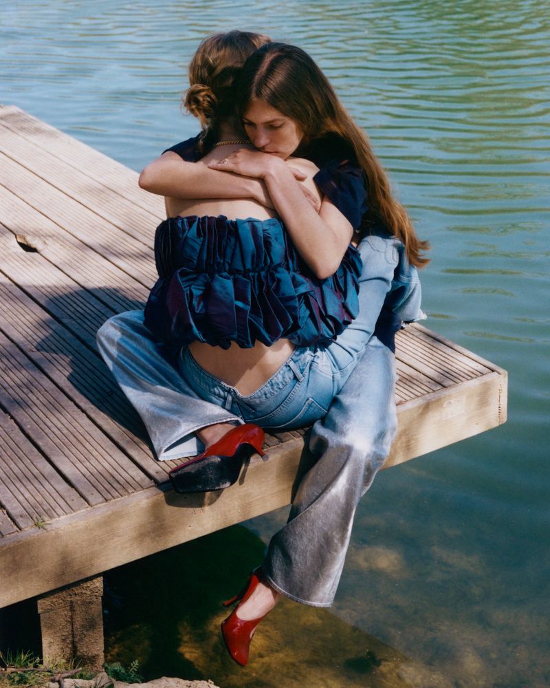 Two women embrace while sitting on a wooden dock by a lake, one wearing high heels and a ruffled top, with sunlight reflecting on the water.