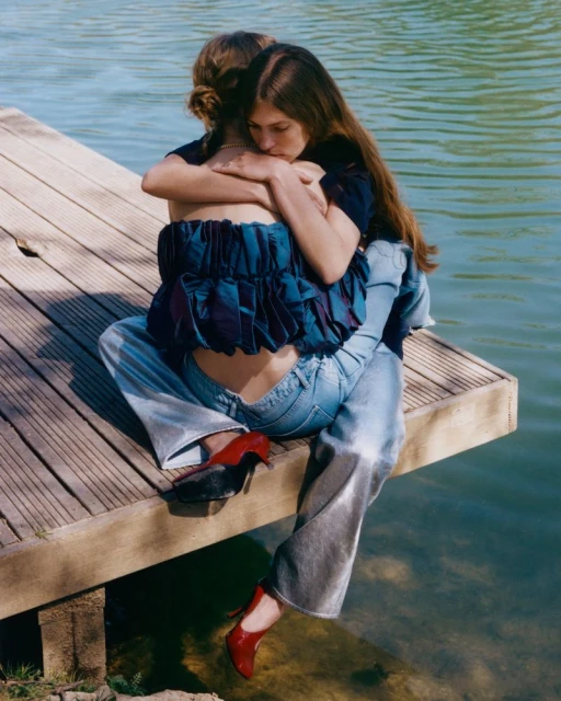 Two women embrace while sitting on a wooden dock by a lake, one wearing high heels and a ruffled top, with sunlight reflecting on the water.