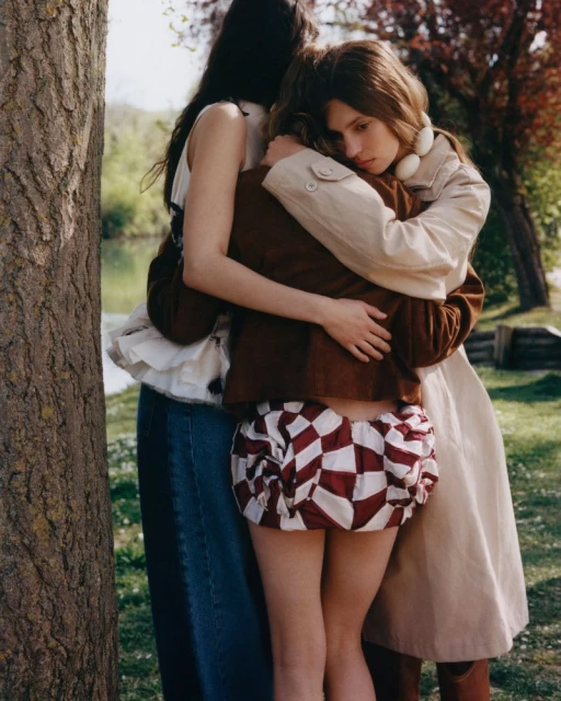 Three people embracing outdoors near a tree. One wears a cream coat, one a brown jacket, and the other a white frilly top and jeans.