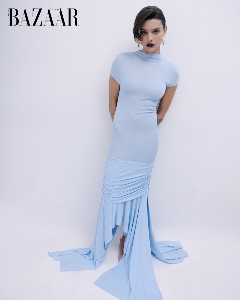 A model poses in a light blue fitted dress with a draped skirt and short sleeves against a plain white backdrop in a studio fashion shot.