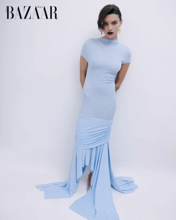A model poses in a light blue fitted dress with a draped skirt and short sleeves against a plain white backdrop in a studio fashion shot.