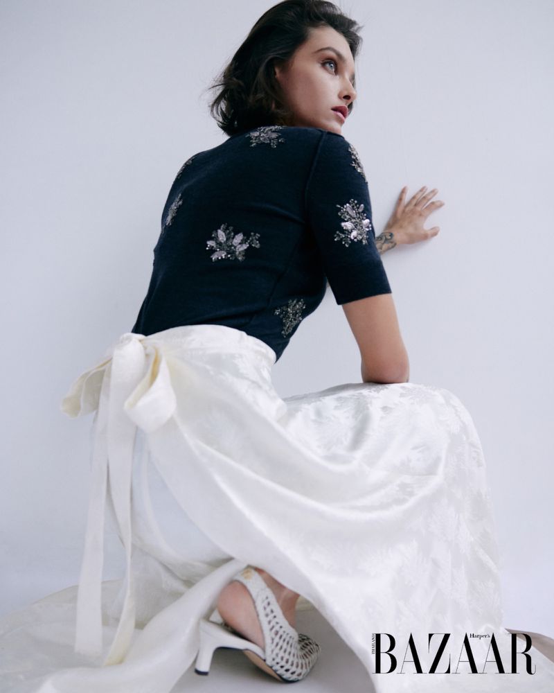 A woman in a black sweater and white skirt poses in a studio with a BAZAAR magazine logo at the bottom of the image.