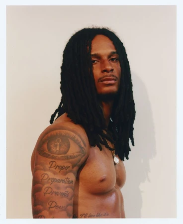 Shirtless Black man with long dreadlocks, arm tattoos, and a gold medallion necklace against a pale backdrop.