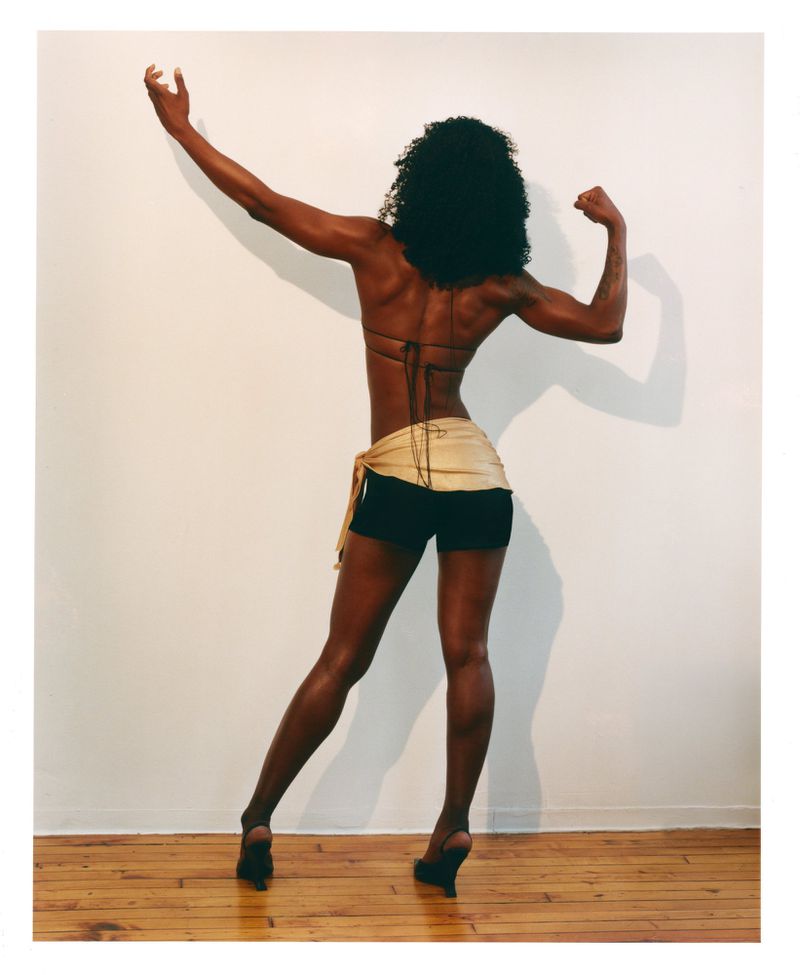 Black woman from behind flexing her arms wearing a string top and dark shorts against a plain white wall.