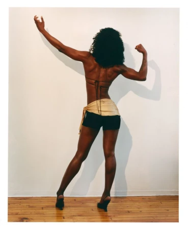 Black woman from behind flexing her arms wearing a string top and dark shorts against a plain white wall.