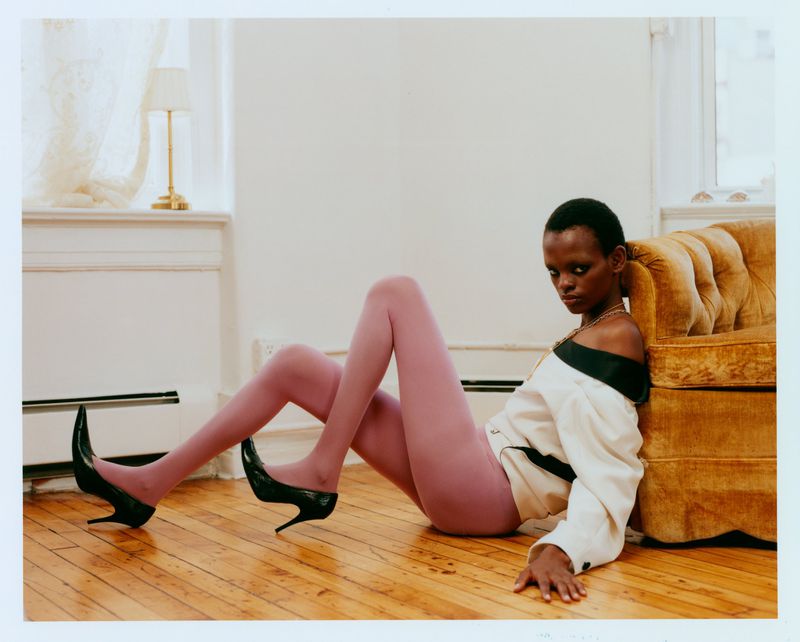 A model wearing mauve tights, an off-the-shoulder cream jacket, and black stilettos reclines on a wooden floor by a tufted yellow sofa.