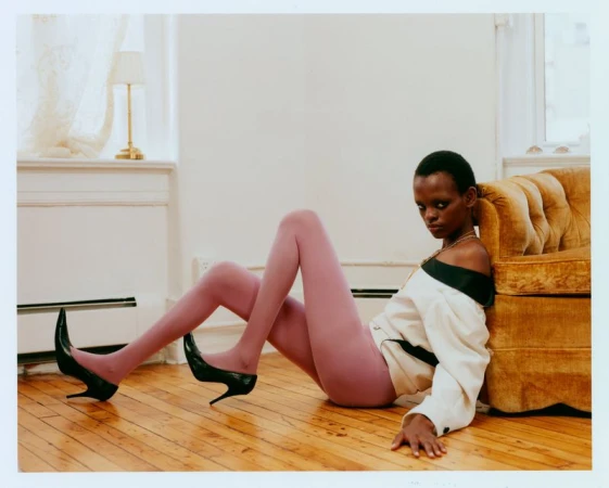 A model wearing mauve tights, an off-the-shoulder cream jacket, and black stilettos reclines on a wooden floor by a tufted yellow sofa.