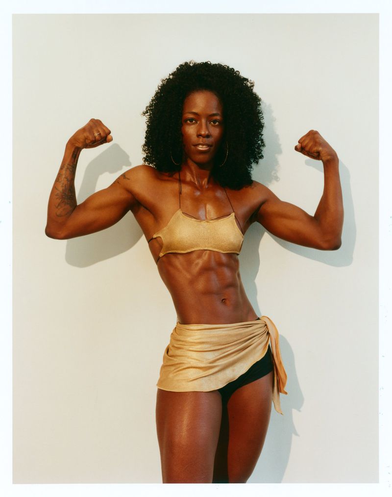 Muscular Black woman flexing both arms while wearing a shimmering gold top and wrap skirt against a light studio backdrop.