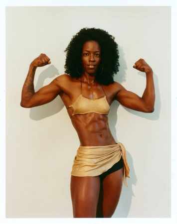 Muscular Black woman flexing both arms while wearing a shimmering gold top and wrap skirt against a light studio backdrop.
