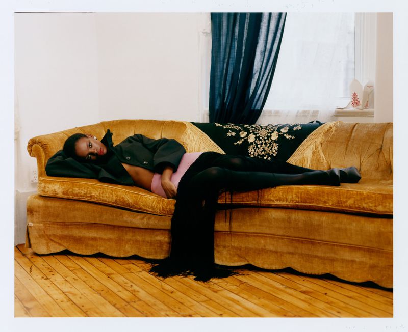 A Black woman in an open dark coat, pink underwear, and black tights reclines across a vintage mustard velvet sofa.
