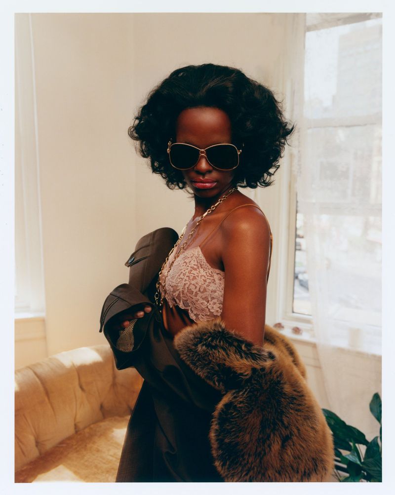 Black woman wearing oversized dark sunglasses, a lace bralette, and a brown coat with fur trim indoors beside a window.