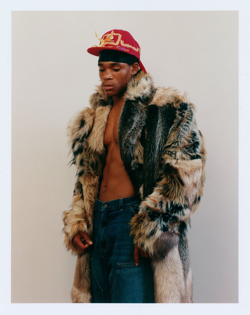 A Black male model posing with an open chest in a voluminous textured fur coat, blue jeans, and a red cap against a pale background.