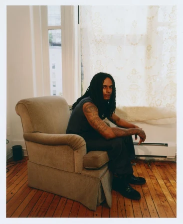 Man with dreadlocks and arm tattoos sitting in a beige armchair on wooden floors beside a window with sheer lace curtains.