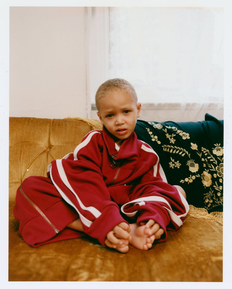 A young child wearing an oversized burgundy tracksuit with white stripes sits barefoot on a mustard velvet sofa.