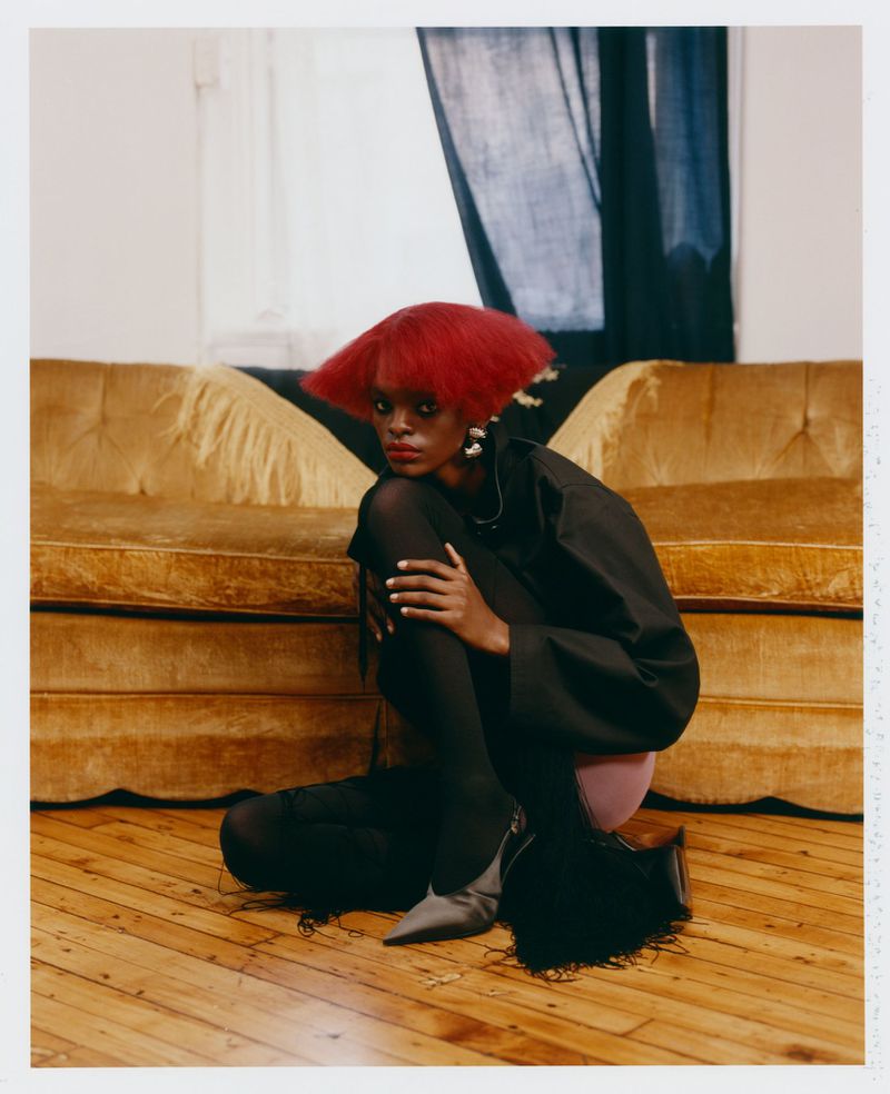 Model with a structured bright red bob haircut crouching on a wooden floor in front of a vintage yellow velvet sofa.