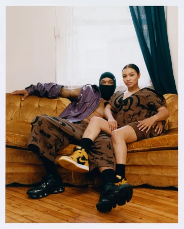 A man wearing a black balaclava and purple jacket seated beside a woman in matching brown printed streetwear on a gold velvet sofa.