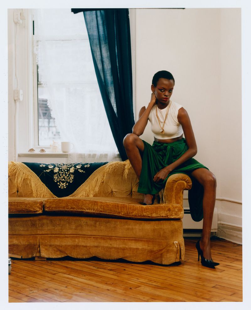 A Black woman seated on the arm of a gold velvet sofa wearing a white crop top, green wrap skirt, and black pointed heels.