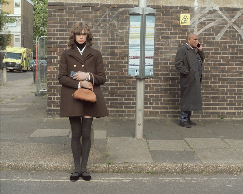 Model standing on a city sidewalk wearing a brown double-breasted coat and knee-high socks, holding a tan handbag near a bus stop.