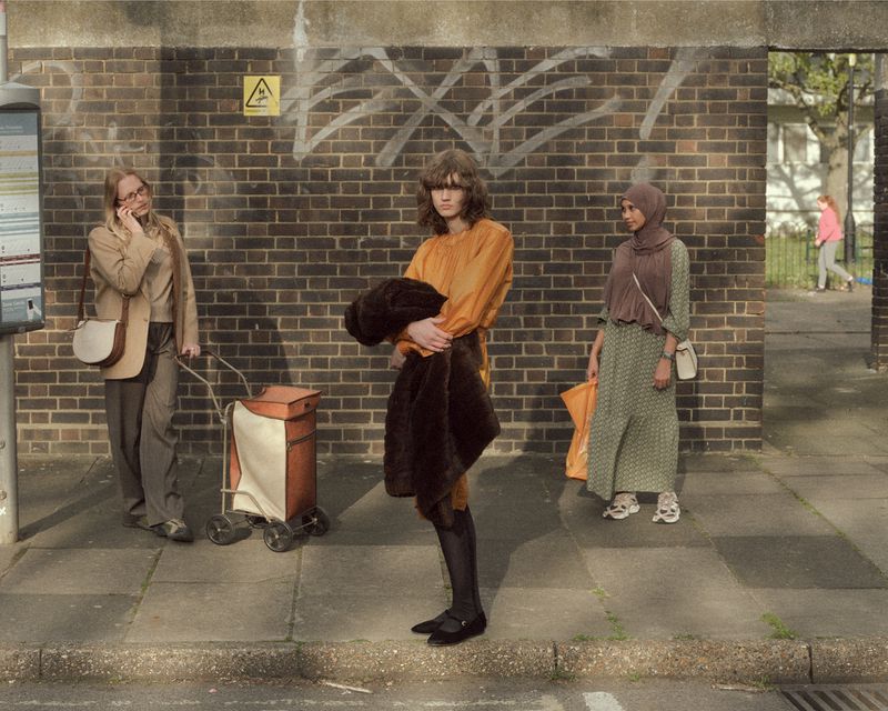 Model Katherine Vinten in an amber tunic dress holding a dark brown faux fur coat, standing on a sidewalk near a brick wall.