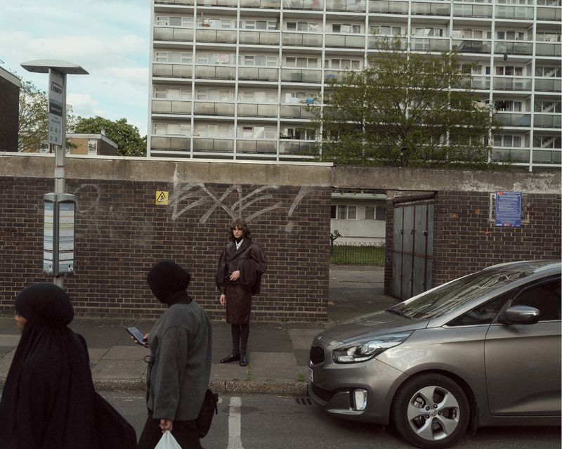 Model standing on a sidewalk in front of a brick wall beneath a large residential tower block in Albion Magazine editorial.