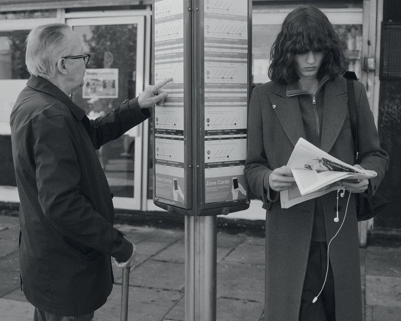 Model Katherine Vinten in a long coat reading a newspaper beside an older man pointing at a bus timetable in black and white.