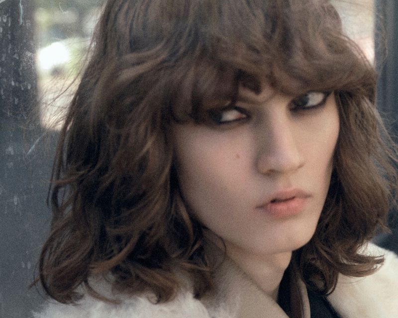 Close-up portrait of a model with dark wavy hair and dark eyeliner wearing a cream-colored textured coat.