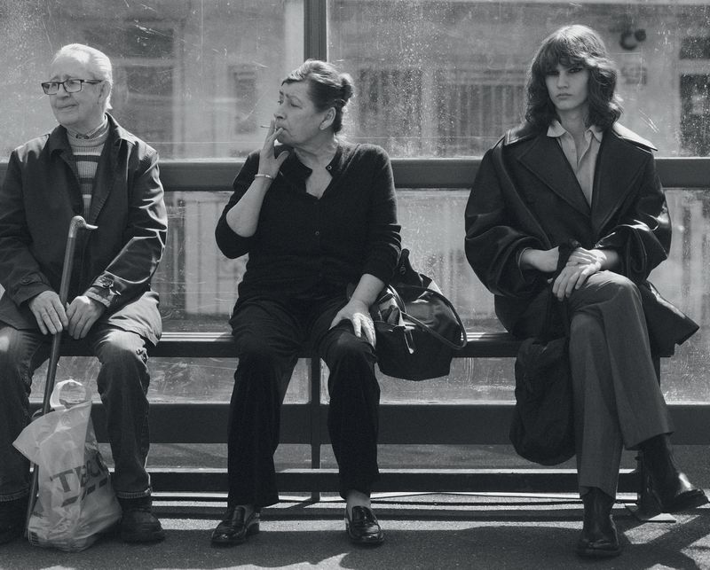 Three people sitting on a bench at a bus stop waiting outdoors in a black and white fashion editorial photograph.