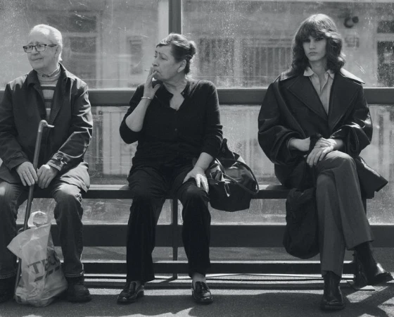 Three people sitting on a bench at a bus stop waiting outdoors in a black and white fashion editorial photograph.